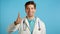 Doctor showing thumb up sign over blue background. Positive young man in medical coat smiles to camera. Winner. Success