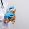 Doctor in a white coat and blue latex gloves holds a brown teddy bear without an eye