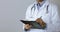 Doctor in white coat writing documents on clipboard on gray background