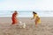 Dog-Friendly Beach. Barefoot Women In Boho Dresses Playing With Cute Puppy On Sandy Coast