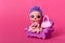 Doll in modern dress and rosy bag sitting on tiny sofa, toy for girl with beautiful big eyes, souvenir for children against rose