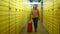 Dolly shot of beautiful Caucasian woman walking with red rolling suitcase in warehouse with yellow lockers. Wide shot