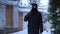 Dolly shot of Caucasian male robber walking with baseball bat on snowy winter backyard. Portrait of confident masked man