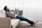 Domestic relax. Young arab guy with wireless headphones and smartphone resting on comfortable sofa at home, side view