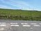 Double lines painted on the side of a country lane with hillside fields surrounded by fences and stone walls with sunlit blue sky