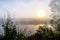 Dramatic mystical twilight landscape with rising sun, tree, reed and fog over water.