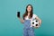 Dreamful girl football fan holding soccer ball, mobile phone with blank empty screen isolated on blue turquoise