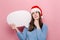 Dreamful young woman holding empty speech bubble stands with fingers crossed, awaits miracle happen, wears Christmas hat