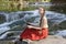 Dreaming beautiful long-haired girl in a red skirt sits with with a laptop on a stone against the backdrop of a cascade of a