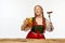 Dreaming redhead woman dressed traditional dirndl, serves appetizing Bavarian sausage on fork and huge beer chole