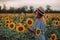 Dreaming young woman in blue dress and hat holding sunflower with a hand in a field of sunflowers at summer, view from her side.