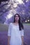 Dreamy image of beautiful woman in white dress walking in street surrounded by purple Jacaranda trees