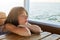 Dreamy little female child leaning on wooden table while having trip on boat, enjoying calm sea, looking with pensive expression i