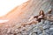 Dreamy lonely brunette hippie in the style of bokho chic sits on gray stones at the sea shore in the rays of the setting sun