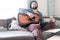 Dreamy man, with eyes closed, plays a guitar, sitting on a soft sofa, next to him is a cat interested in what is