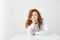 Dreamy tender young girl with red curly hair thinking dreaming sitting at table over white background.