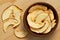 Dried apple slices in a brown wood bowl next to spilled dried apple slices on light wood background from above