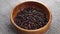 Dried black pepper grains fall into a wooden bowl on a jute burlap