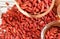 Dried goji berries in wooden bowls, more scattered over white boards table under, closeup detail from above