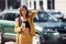 With drink in hands. Young fashionable woman in burgundy colored coat at daytime with her car