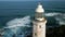 Drone circles historic Dondra lighthouse in Sri Lanka, white beacon in vibrant blue ocean, greenery. Waves crash against