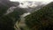 The drone flies over a narrow mountain river in lowland between mountains and pine forest. Foggy aerial view.