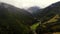 The drone flies over a narrow mountain road in green lowland between mountains and pine forest. Foggy aerial view.