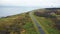 Drone flying above black car moving along clear road near beautiful lighthouse sea bay and autumn field on cloudy day.