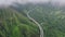 Drone flying through clouds above epic highway winding along green mountain 4K