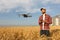 Drone hovers in front of farmer with remote controller in hands near grain elevator. Quadcopter flies near pilot