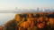 Drone moving to beautiful sunrise city skyline panorama over still lake waters and sunny yellow autumn forest.