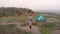 Drone shot, mother and child on hike standing by tent on top of hill. Little girl hiker standing in nature arms