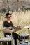 A drummer in glasses plays drums in the countryside. The man has a black hat, behind him an old castle