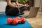 Dumbbells and unrecognizable sporty woman making meditation yoga fitness stretching at home via laptop. Young woman losing weight