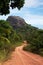 Dusty road going through green colored bushes leading to a big black rock.