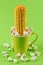 An ear of corn sticks out of a green cup, a symbol of fertility, surrounded by scattered popcorn, concept