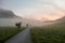 Early morning haze in the Alps. There is a narrow road leading to high mountains through a meadow. The high Alpine peaks