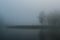 Early summer morning fog on river. Island, overgrown with wood, barely visible in dense fog. Thick morning mist brings depressive