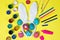 Easter concept flat lay: bunny ears, painted eggs, paints, felt-tip pens and markers on a yellow background.