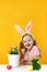 Easter concept. A little girl sits at a table with a potted plant, eggs and a rabbit. Happy child in bunny costume yellow