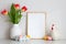 Easter home decorations on table. Picture frame mockup, decorative chicken, Easter eggs, vase of tulips on white wall background