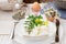 Easter table setting, white plates, cutlery, napkin, flowers in eggshell, green twigs, crystal cup, basket