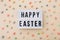 Easter wishes on lightbox with chocolate mini eggs scattered on a beige background, top view