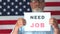 Economic crisis. Depressed man with a beard is holding a sign saying need job. American flag on the background