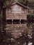 An eerie looking empty boat shack surrounded by trees and ferns with reflection of the shack on the water