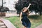 Effective girl with long hair in black dress is posing to camera in city park. She holds hand above and looks excited