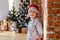 An eight year old boy in pajamas and a Santa hat stands near a brick wall against the background of a Christmas tree