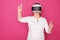 Elderly in 60s woman plays in VR helmet. She stands in white blouse on pink background and looks happy