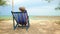 An elderly Asian woman in a hat sits on a deck chair by the beach.