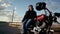 An elderly biker in a leather jacket and gloves comes to the bike on the background of an empty road and sits on it. The